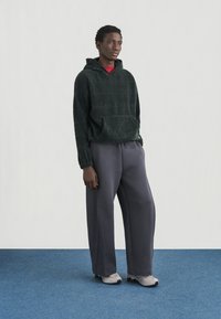 Σκούφος από πράσινο fleece με τσέπη καγκουρό, σε συνδυασμό με χαλαρό γκρι παντελόνι φόρμας και ανοιχτό γκρι αθλητικά παπούτσια, σε φόντο μπλε χαλιού.