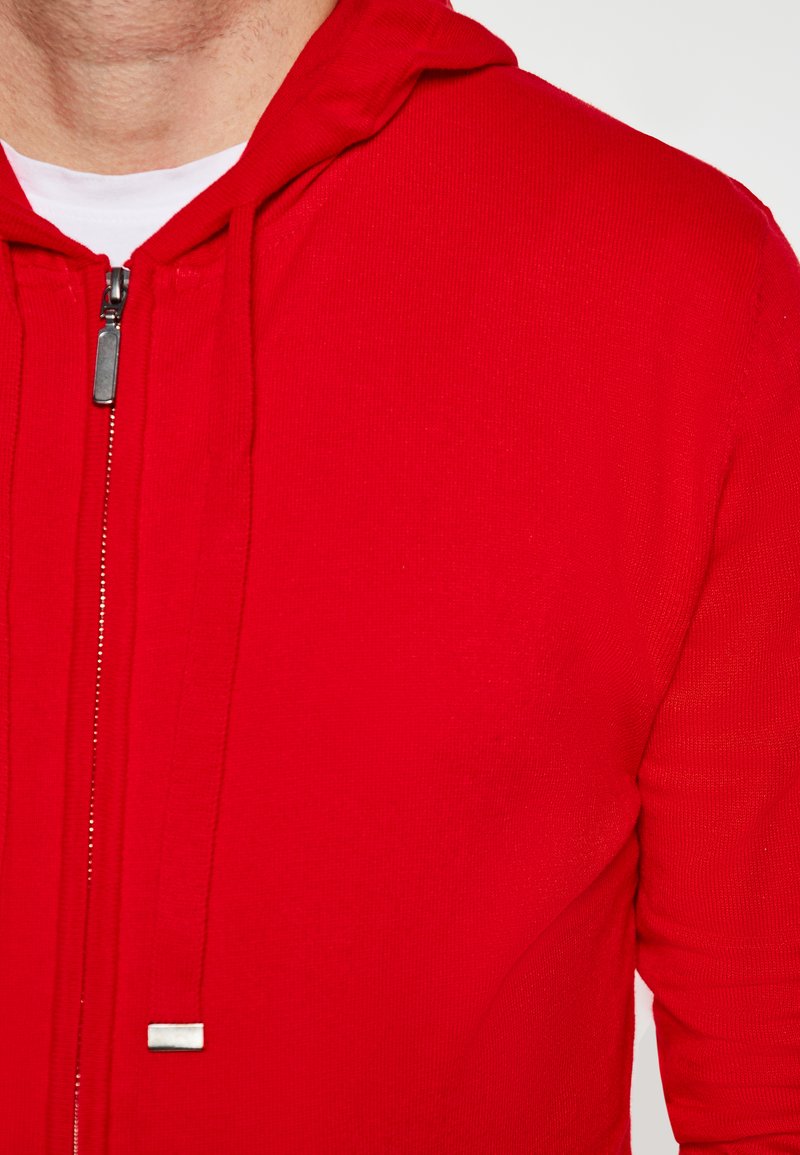 Felpa rossa con zip, caratterizzata da un tessuto testurizzato, cappuccio e dettagli in metallo. Design semplice senza ulteriori motivi o accentuazioni.
