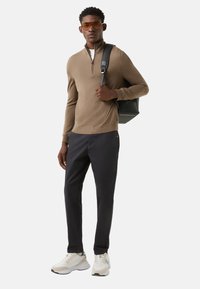 Braune Sweatjacke mit Reißverschluss, dunkle Hose und weiße Turnschuhe. Accessoires umfassen eine schwarze Tasche und orange getönte Sonnenbrille.