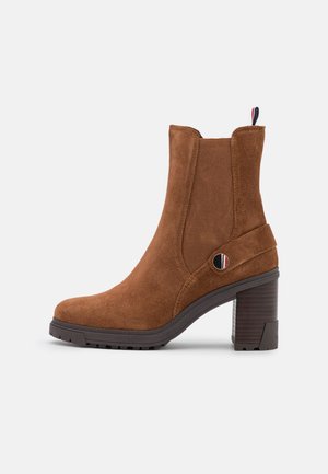 Botte de cheville en daim marron avec un talon bloc, des panneaux latéraux élastiques, une languette rayée et un accent en métal circulaire sur le côté.