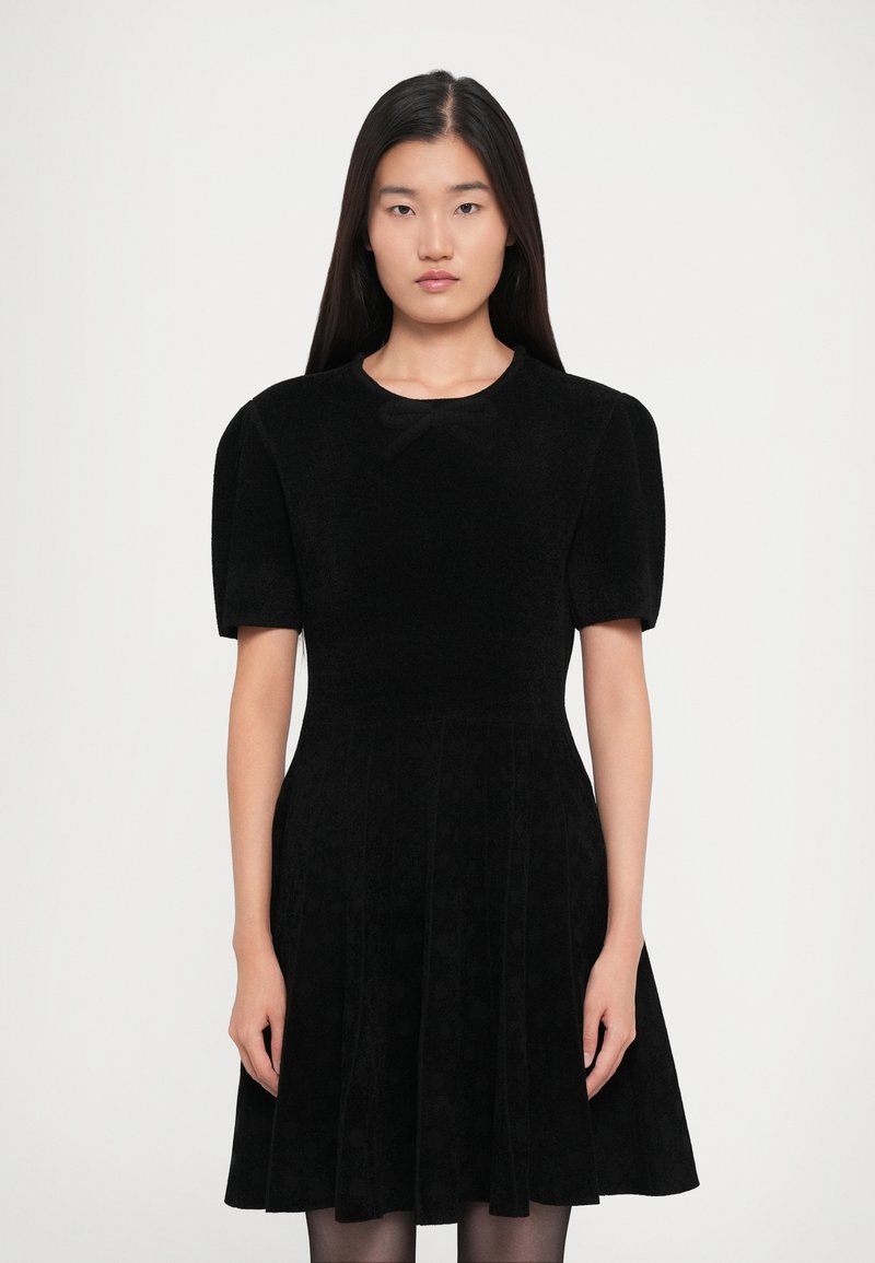 Woman with long black hair wearing a black short-sleeve dress with a bow at the neckline against a plain white background.
