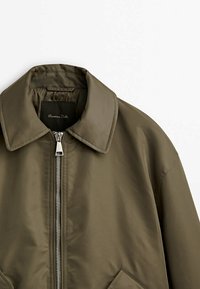 Veste zippée marron-vert avec col pointu et poches diagonales, dotée d'un tissu lisse et de détails de surpiqûres visibles.