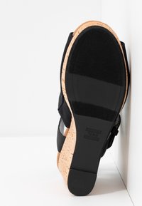 Sandale compensée noire avec semelle en liège, bande en caoutchouc et bride à la cheville, semelle affichant le nom de la marque TOMS sur fond blanc.