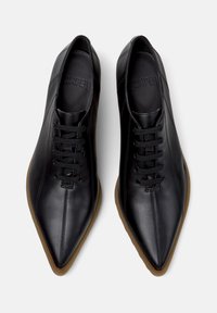 Μαύρα δερμάτινα παπούτσια με μυτερή μύτη και κορδόνια. Η σόλα είναι καφέ από καουτσούκ, προσφέροντας μια αντίθετη λεπτομέρεια στο πάνω μέρος.