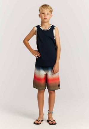 Blond jongen staat met een hand op zijn heup, draagt een marineblauw mouwloos shirt, meerkleurige geometrische korte broek, slippers en een enkelbandje aan zijn rechterbeen.