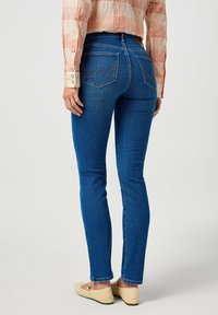 Jeans attillati blu in denim, con due tasche posteriori dal design a zigzag, abbinati a scarpe beige piatte.