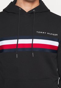 Haut à capuche noir avec une texture tricotée circulaire, présentant un motif à rayures horizontales en marine, blanc et rouge. Logo imprimé en blanc sur la poitrine.