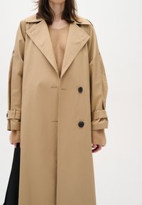 Beige trench coat with a double-breasted design, black buttons, and oversized sleeves. Worn over a light brown sweater.