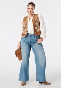 Bruine suede vest met bloem borduurwerk, witte textuur blouse, lichtblauwe flared jeans en beige enkellaarzen; houdt een bruin hoed vast.