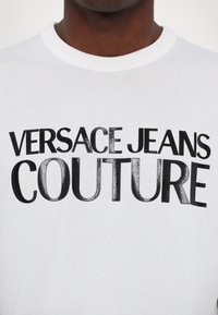 Biała bawełniana koszulka z odważnym czarnym nadrukiem: "VERSACE JEANS COUTURE". Klasyczny okrągły dekolt i krótkie rękawy.