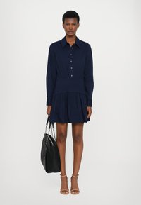 Abito blu navy con bottoni, maniche lunghe, vita aderente, gonna svasata e pieghe sottili. Abbinato a una borsa nera e sandali color carne.