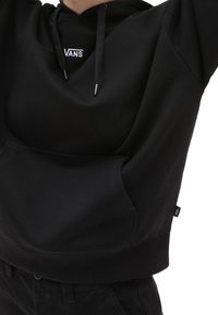 Felpa corta nera con tasca frontale, presenta un piccolo logo bianco "VANS" sul petto, realizzata in tessuto liscio con una texture morbida.