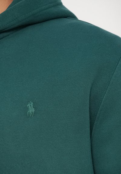 Kadar izbliza tamnozelene tkanine s toniranim izvezenim logom igrača polo na šavu ramena odjevnog predmeta.