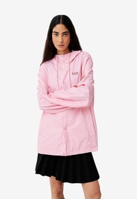 Helle rosa wasserfeste Jacke mit Kapuze, Druckknöpfen und kleinem Logopatch. Darunter wird ein schwarzer Plisseerock getragen.