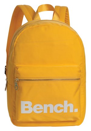 Gelber Stoff-Rucksack mit runder Form, einer Frontreißverschlusstasche und großem weißen "Bench."-Logo. Verfügt über verstellbare Träger und eine glatte Oberfläche.