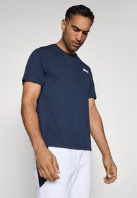 Sötétkék pamut póló rövid ujjal, kerek nyakkal és egy kis logós ruhával a bal mellrészen. Sima textúra, laza fazon.