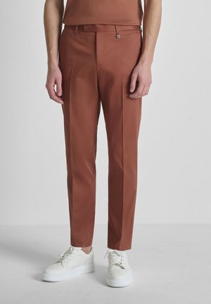 Hombre vestido con pantalones cónicos color óxido y zapatillas blancas, de pie frente a un fondo liso de color gris claro.