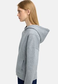 Szara bluza z kapturem wykonana z miękkiego materiału, z przednim suwakem, bocznymi kieszeniami oraz luźnym krojem z ściągaczami przy mankietach i dolnej krawędzi.