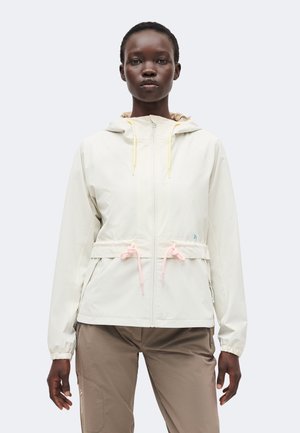 Kobieta w jasnobeżowej kurtce z kapturem, zapinanej na zamek błyskawiczny z przodu i różowymi sznurkami, w połączeniu z taupe spodniami, stojąca na jednolitym tle.