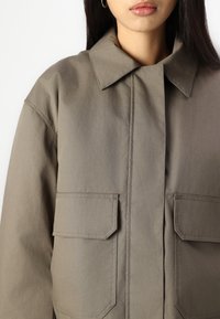 Giacca khaki con bottoni, caratterizzata da un colletto ampio, due tasche frontali e una vestibilità rilassata. Il tessuto appare liscio con una leggera lucentezza.