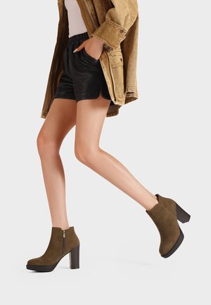 Persona che cammina indossando stivaletti in camoscio verde oliva con tacco a blocco, pantaloncini neri, top bianco e giacca marrone.