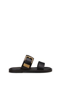 Schwarze flache Sandale mit zwei Riemen, einer mit einer großen goldenen Schnalle und einer kleinen goldenen Markenplakette, vor weißem Hintergrund.