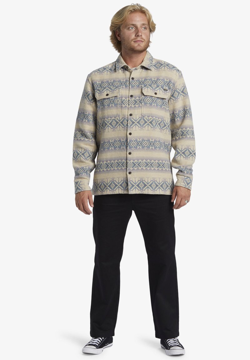 Camisa de manga larga, con botones, en beige con patrones geométricos en azul. Presenta dos bolsillos en el pecho y un corte regular. Combinada con pantalones negros y zapatillas.