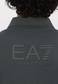 Tamsiai pilkas polo marškinėliai su tekstūruota gumine nugaros logotipu "EA7 EMPORIO ARMANI." Klasikinio tipo apykaklė su subtiliais siūlėmis.
