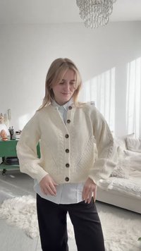 Νεαρή γυναίκα προσαρμόζει το κρεμ πλεκτό κασκόλ με μεγάλα κουμπιά, στέκεται σε φωτεινό καθιστικό με πράσινη ντουλάπα, λευκό καναπέ και πολυέλαιο.