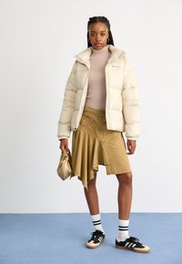 Jachetă puffer crem, pulover bej ajustat cu guler înalt, fustă asimetrică kaki, adidași negri și bej, ținând o geantă de mână aurie, șosete albe cu dungi negre.