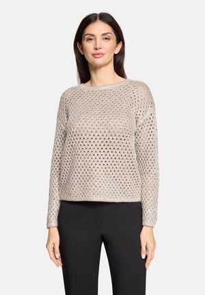 Vrouw draagt een beige grof gebreide trui met lange mouwen en zwarte broek, staand tegen een effen lichte achtergrond.