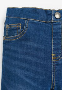 Tumedat sinised denim püksid klassikalise lõikega. Omavad kollast tikandit, eesmist taskut ja metallist nööbi riistvara vööl.
