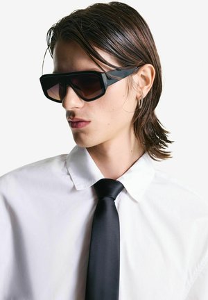 Occhiali da sole neri rettangolari con lenti sfumate, indossati con una camicia bianca col colletto e una cravatta nera. Design elegante con hardware minimale.