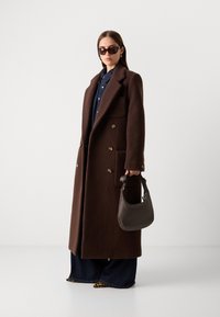 Cappotto in lana marrone con design doppiopetto, grandi tasche e bottoni dorati. Abbinato a una camicia di jeans, jeans a gamba larga e una borsa scura.