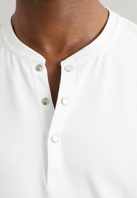 Eine dunkelhäutige Person trägt ein weißes, strukturiertes Henley-Shirt mit drei metallenen Druckknöpfen, die am Kragen teilweise geschlossen sind.