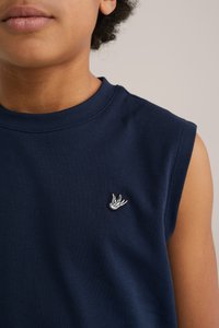 Temno modra brezrokavnica z okroglo izrezom in mehko bombažno teksturo, ki ima na prsih majhen beli vezen ptičji logotip.