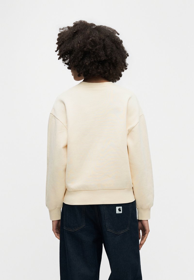 Hellbeiger Pullover mit Rundhalsausschnitt, Raglanärmeln und geripptem Saum, kombiniert mit dunkler Denim-Hose mit Logo-Patch.