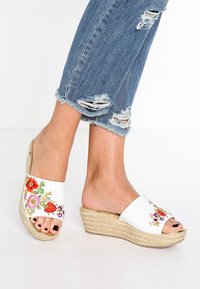 Sandales compensées blanches avec un motif floral brodé, dotées d'une plateforme en corde naturelle et d'un bout ouvert, associées à un jean déchiré.
