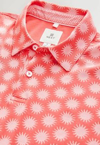 Camisa polo color coral con un patrón de sol en rosa claro. Diseño con cuello y tres botones, tela suave y cuello interior con patrón.