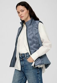 Gepolsterte graue Weste mit hohem Kragen und Frontreißverschluss, die über einem weißen Hemd und blauen Jeans getragen wird. Die Weste hat eine glatte, glänzende Textur.