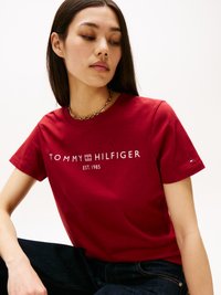 Camiseta de algodón roja con un logo blanco que dice "TOMMY HILFIGER EST. 1985." Mangas cortas y un cuello redondo, fabricada con tejido texturizado.