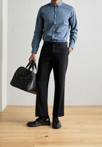 Niebieska koszula z długim rękawem zapinana na guziki, czarne spodnie i czarne skórzane buty. Nosi zwartą czarną skórzaną torbę z zaokrąglonymi krawędziami.