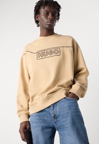 Beige pulóver fekete "HUGO" logóval. Kerek nyakú, hosszú ujjú, bordázott mandzsettás. Világoskék farmernadrággal párosítva.