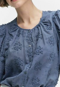 Ljusblå blus med blommönsterbroderi, korta puffärmar, rund halsringning och knäppning på framsidan. Mjuk textilstruktur.