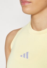 Világos sárga, ujjatlan felső magas nyakkivágással és lila Adidas logóval. Az anyag simának és testhezállónak tűnik, hangsúlyozva a vállak területét.