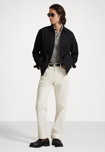 Black jacket, gray polo, and cream pants. Model wears sunglasses with black shoes. Jacket features a zipped front and pockets.