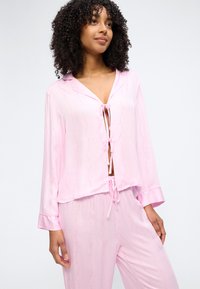Γυναίκα που φορά ελαφρύ ροζ σατέν σετ πιτζάμες με μακριά μανίκια και δέσιμο στο πάνω μέρος και στο παντελόνι.