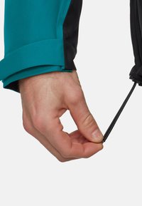 Hånd justerer sort elastisk snor på ærmet af en blå og sort jakke mod en hvid baggrund.