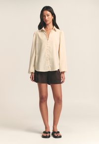 Cream button-up blouse with long sleeves, shaped hem, and soft texture, paired with brown shorts and black flat sandals.