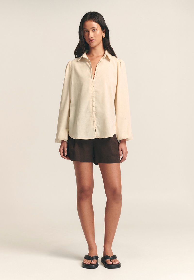 Cream button-up blouse with long sleeves, shaped hem, and soft texture, paired with brown shorts and black flat sandals.